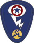 Oval shaped shoulder patch with a deep blue background. At the top is a red circle and blue star, the patch of the Army Service Forces. It is surrounded by a white oval, representing a mushroom cloud. Below it is a white lightning bolt cracking a yellow circle, representing an atom.