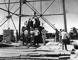 Men stand around a large oil-rig type structure. a large round object is being hoisted up.