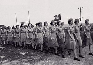 A group of women in uniform marching. They carry a small flag with the Corps of Engineers; castles surmounted by the letter D.